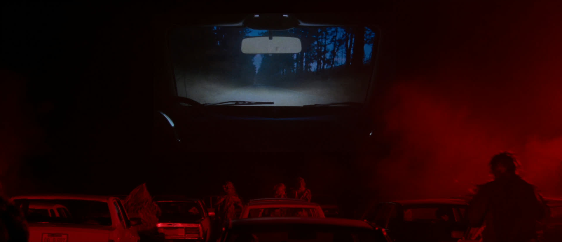 Lo schermo del drive-in con la strada nel bosco, le auto ferme e figure tra le auto